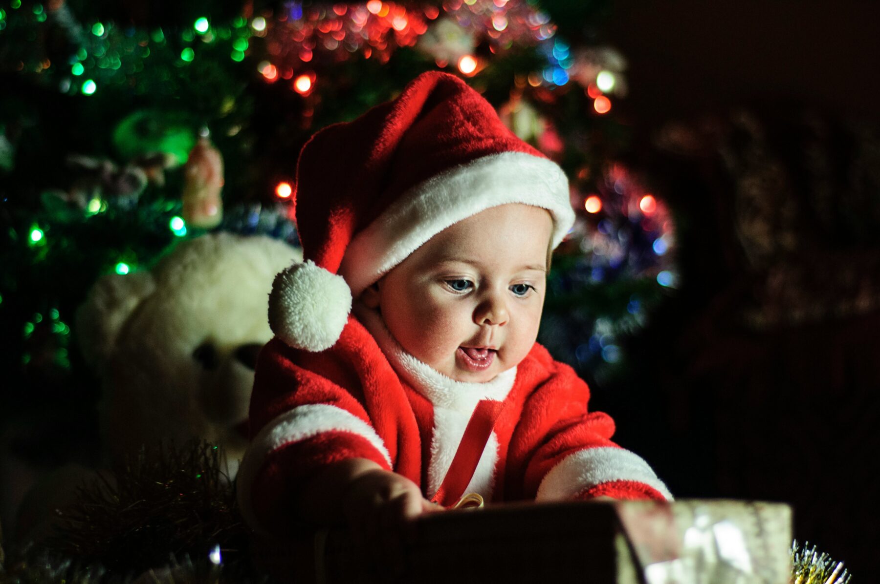Baby in a red Santa outfit smiling and reaching for a gift box in front of a Christmas tree, perfect for DIY Christmas baby photo ideas at home.